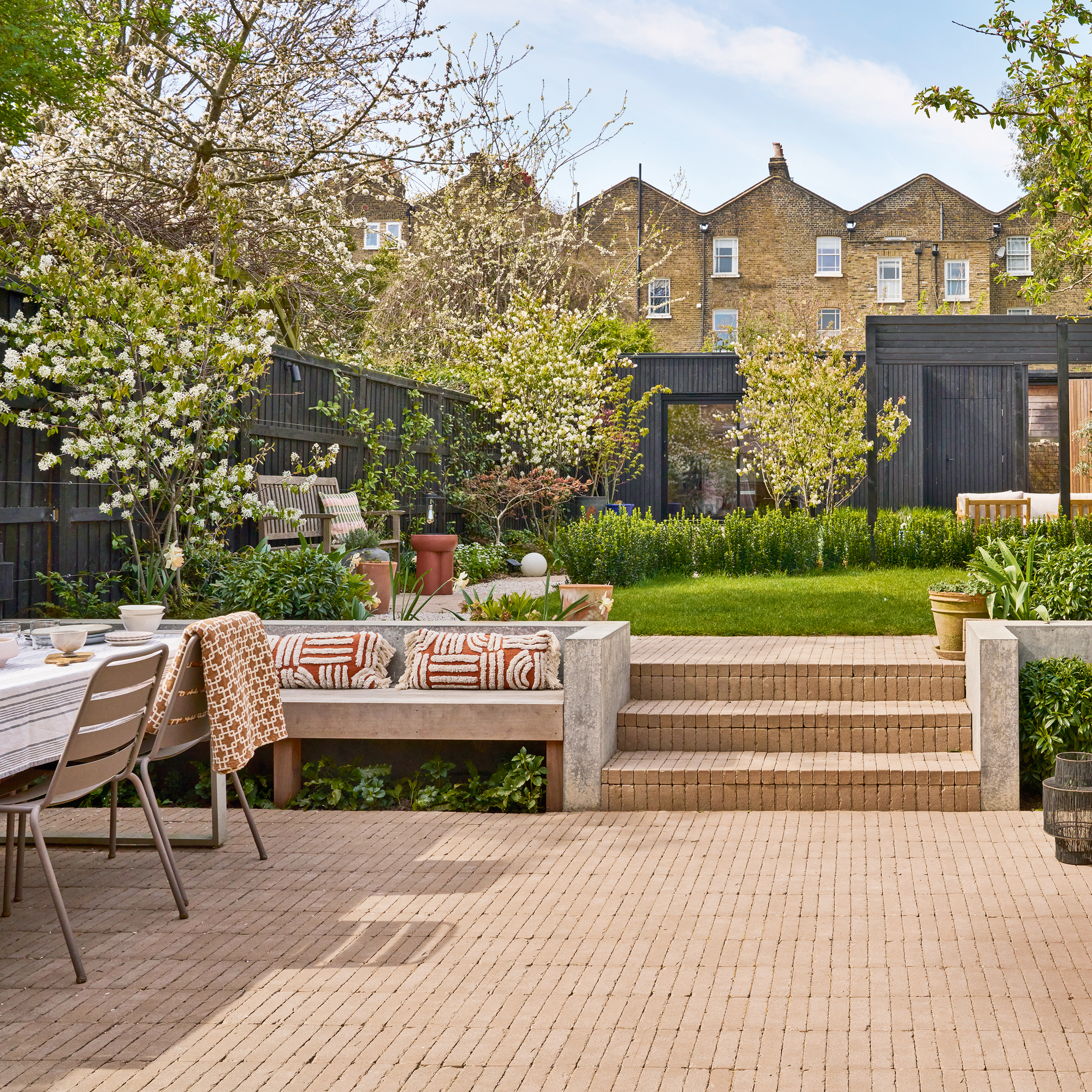 A large back garden with a black time studio at the end, trees and lawn in the middle and a brick terrace in the foreground with a dining area