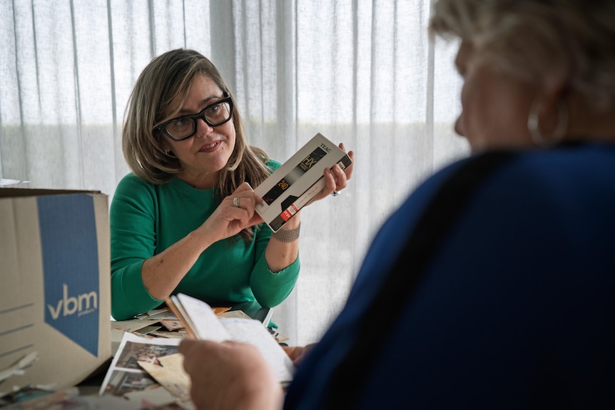 Lissanne Oliver, who has long hair and glasses, holds a VHS band while looking at Sue.