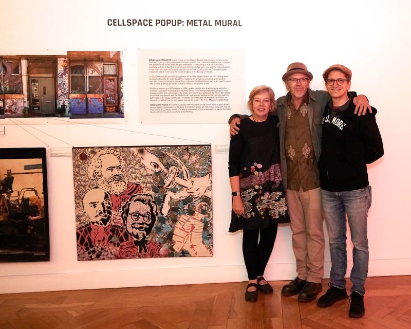 Three people stand together in front of a gallery wall in which works of art and a sign read "Zellspace Popup: Metal Mural.
