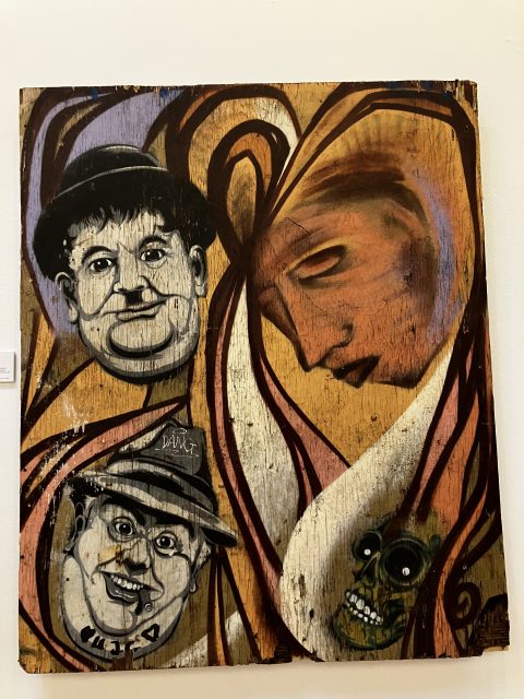A painting over wood with abstract swirls, a large melancholy face in the profile, two cartoons of male faces and a green skull.