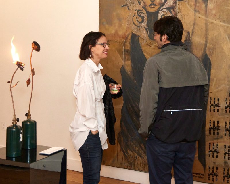 Two people stand in front of a large work of art in a gallery. You keep a drink. A metal rose in Flame, one of the works of art in the gallery, is visible nearby.