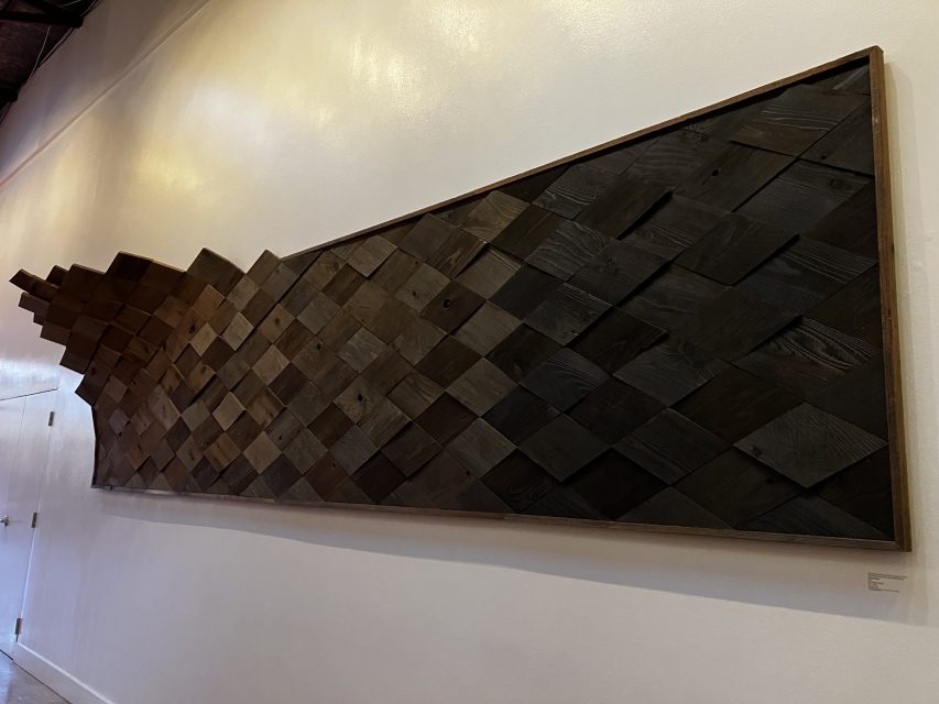 A work of art mounted on the wall consists of dark wooden tiles that are arranged in a geometric, gradually wavy pattern within a rectangular frame.