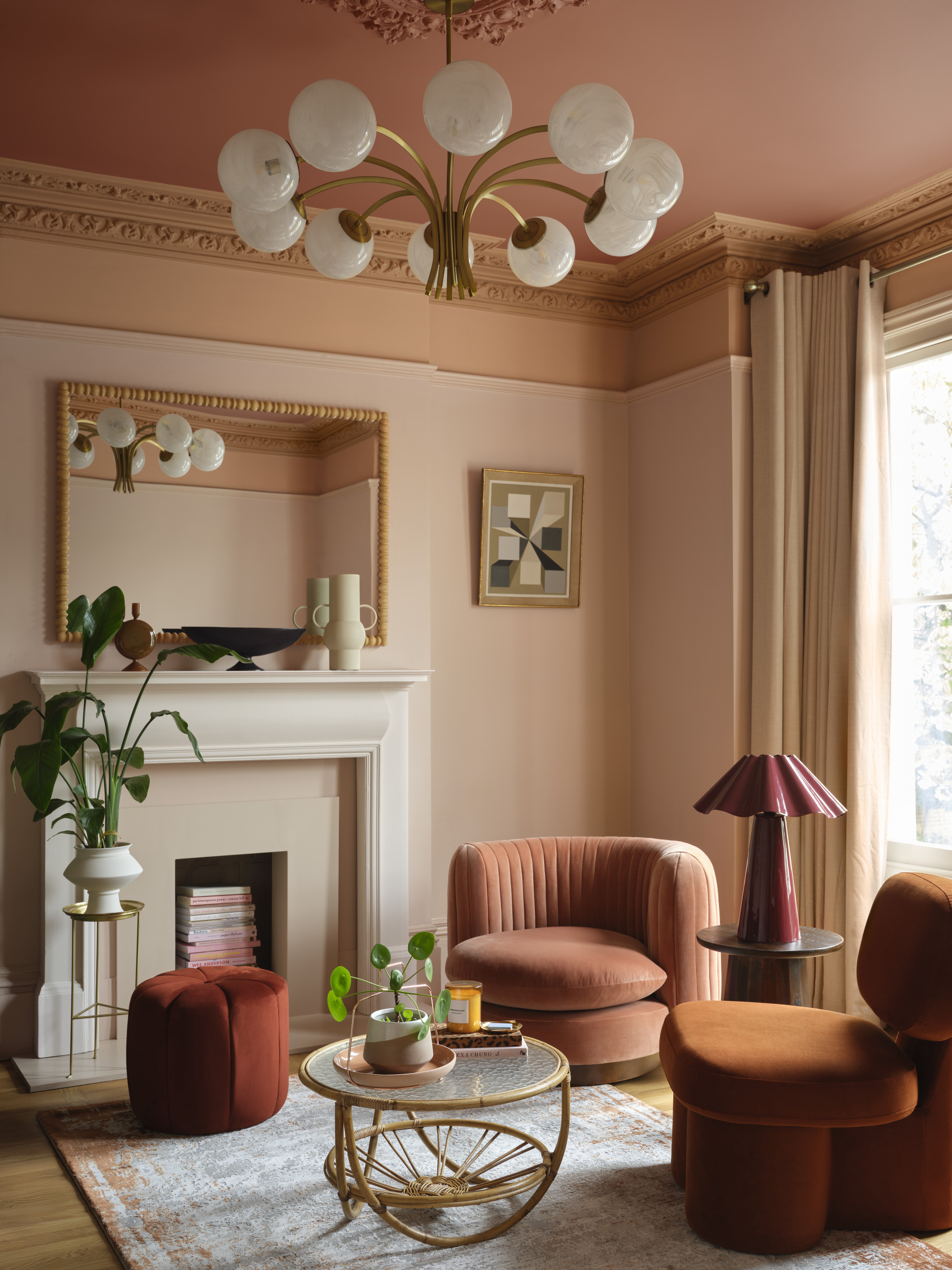 A living room painting in a color cap technology with different colors peach and pink colors. There are crown shapes around the ceiling and a white lamp lamp. There are two pink and orange -colored, velvety akent chairs and a fire space in the room.