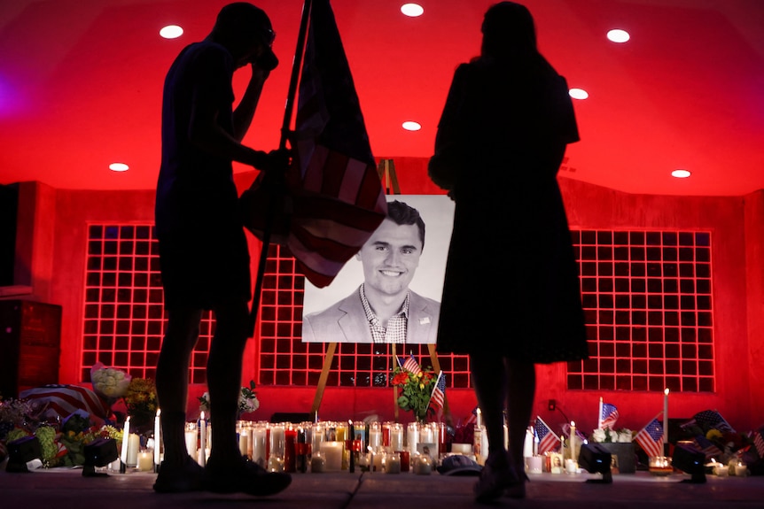 Silhoute of mourners can be seen in front of candles that are placed under a large picture by Charlie Kirk.