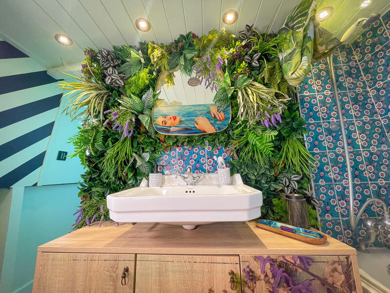 A bathroom with a white sink on a wooden cabinet, framed by a wall of works of art and a mirror that represents the face of a woman.