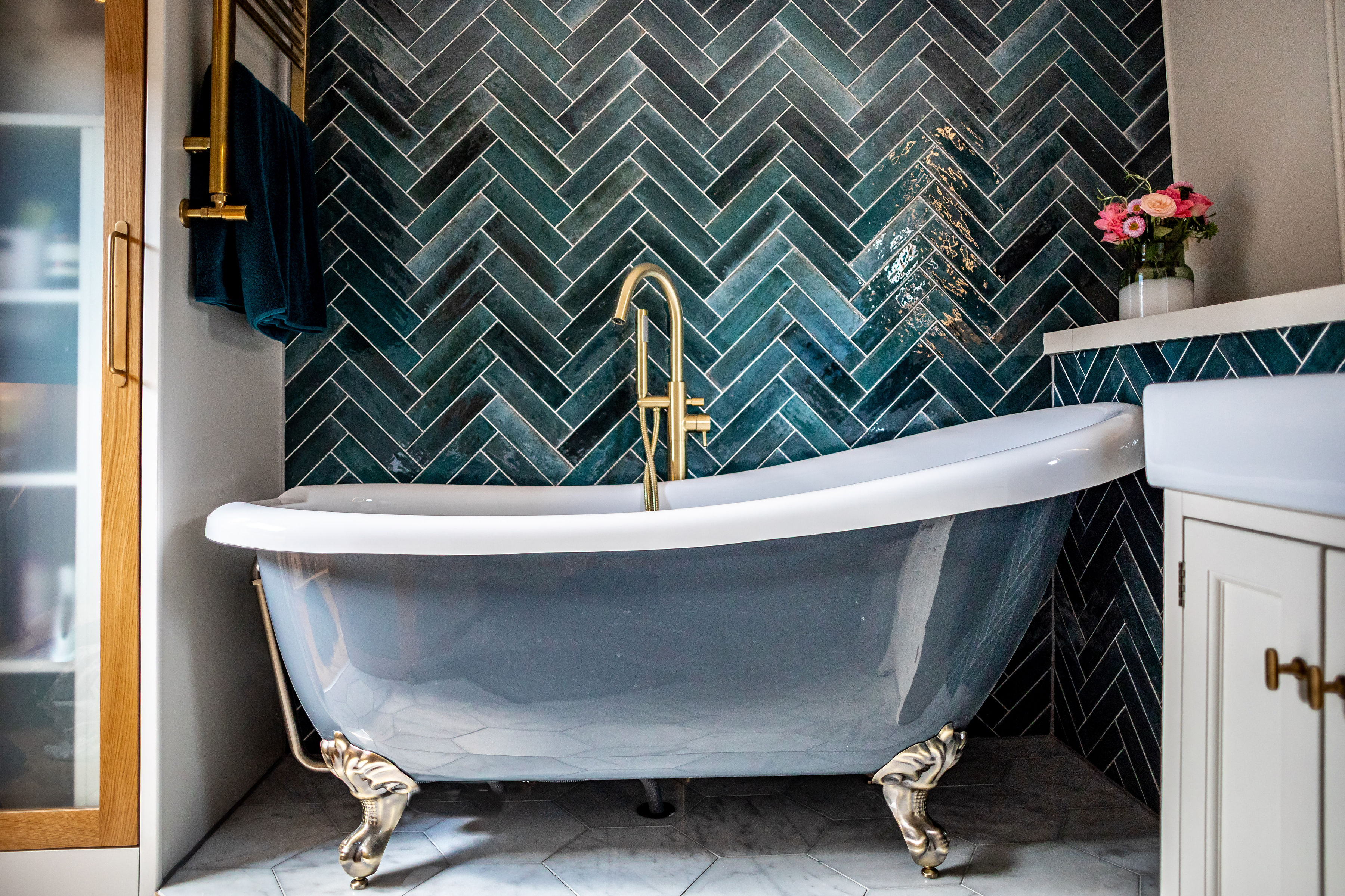 A modern bathroom with a Clawfoot bathtub with a brass fitting that plays against a dark blue -green herringbone tile.