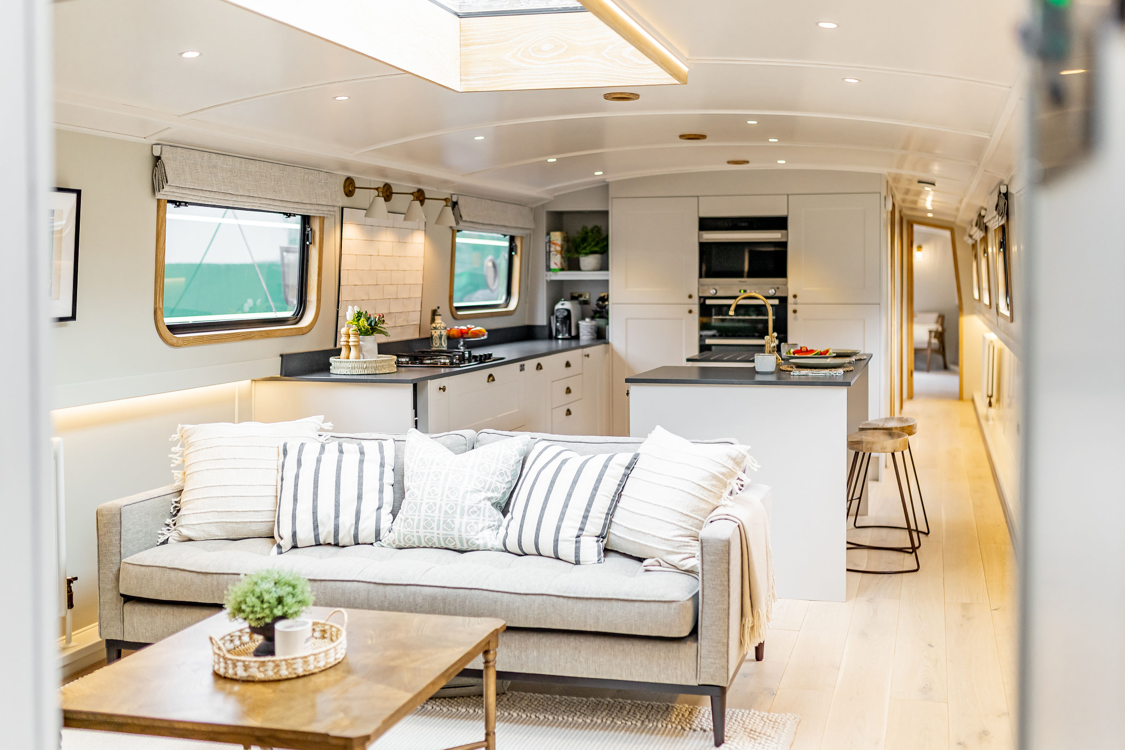 Living area and kitchen of a narrow boat with light walls, couch, kitchen cabinets and wooden floors.