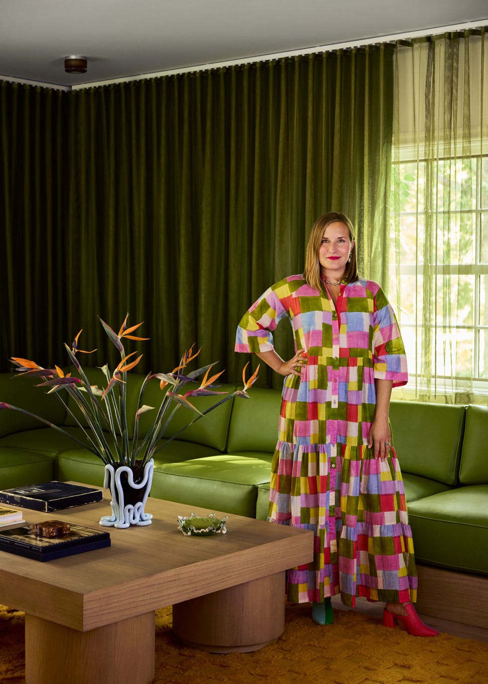 A woman who is dressed in a colorful patterned dress is confident in a stylish interior with a green couch and decorative elements.