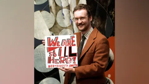 Anderson and Garland An auctioneer in the air holds a work of art that consists of the words "We are still here!" In red and black against a white background. It has dark hair, a narrow, shortened beard and wears glasses and a brown jacket over a brown and white shirt and a dark tie.