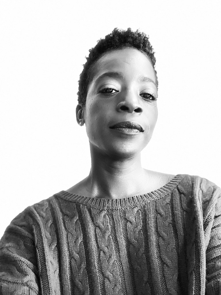 A black and white headshot of the artist wearing a cable knit sweater. She has short dark hair and looks into the camera with a slight smile. The background is bright white.