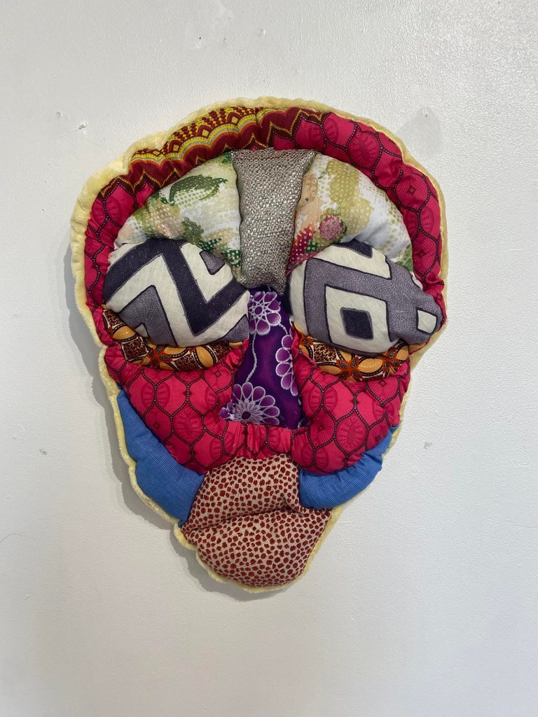 A wall hanging soft sculpture of a face made of fabric with different patterns: in blue, red, gold, green and dark blue.