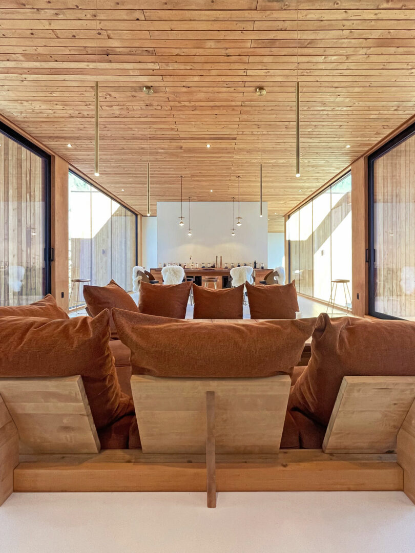 A modern living room with wooden ceiling and walls, large brown upholstered chairs, pendant lights and a dining area in the background with lots of natural light.