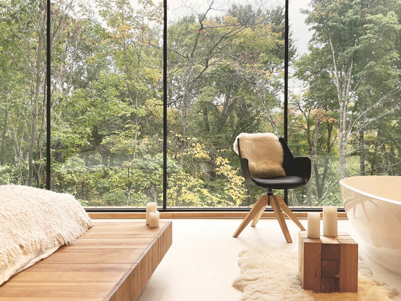 A modern bathroom with large windows overlooking the forest, a wooden platform, a black chair with a fur throw, a white bathtub and candles.