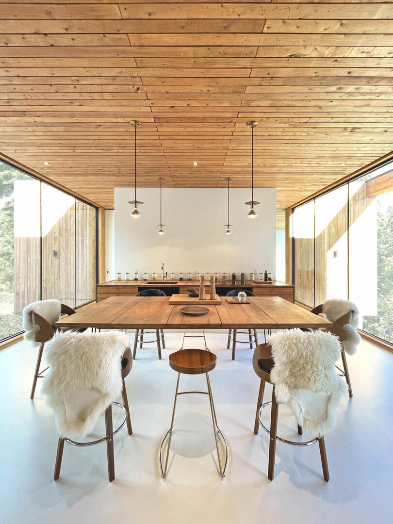 A modern dining room with a large wooden table, minimalist decor, pendant lights and floor-to-ceiling windows; White fur throws are draped over the chairs.