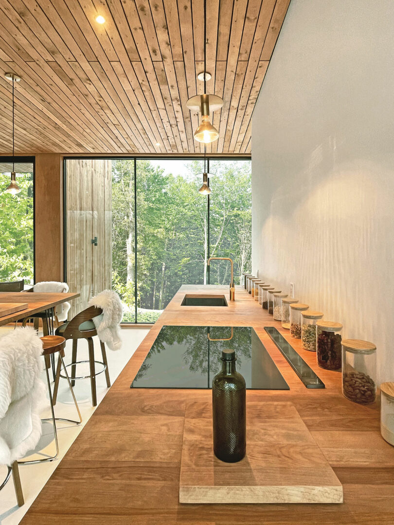 Modern kitchen with wooden countertops, built-in hob, dry goods jars and large windows overlooking a wooded area.