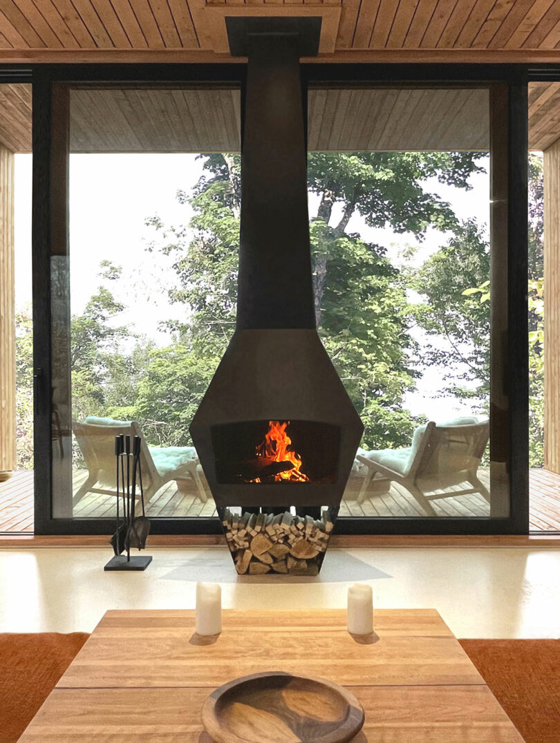 A modern black fireplace with a burning fire stands in front of large glass windows overlooking a patio with lounge chairs and trees outside. In the foreground is a wooden coffee table.