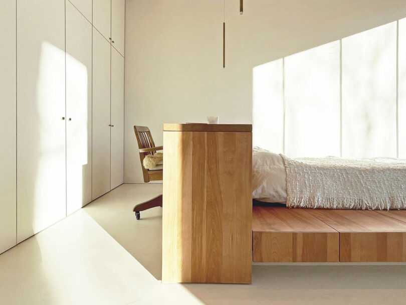 Minimalist bedroom with white walls, built-in wardrobes, a wooden bed platform, a wooden chair and sunlight streaming through large windows.