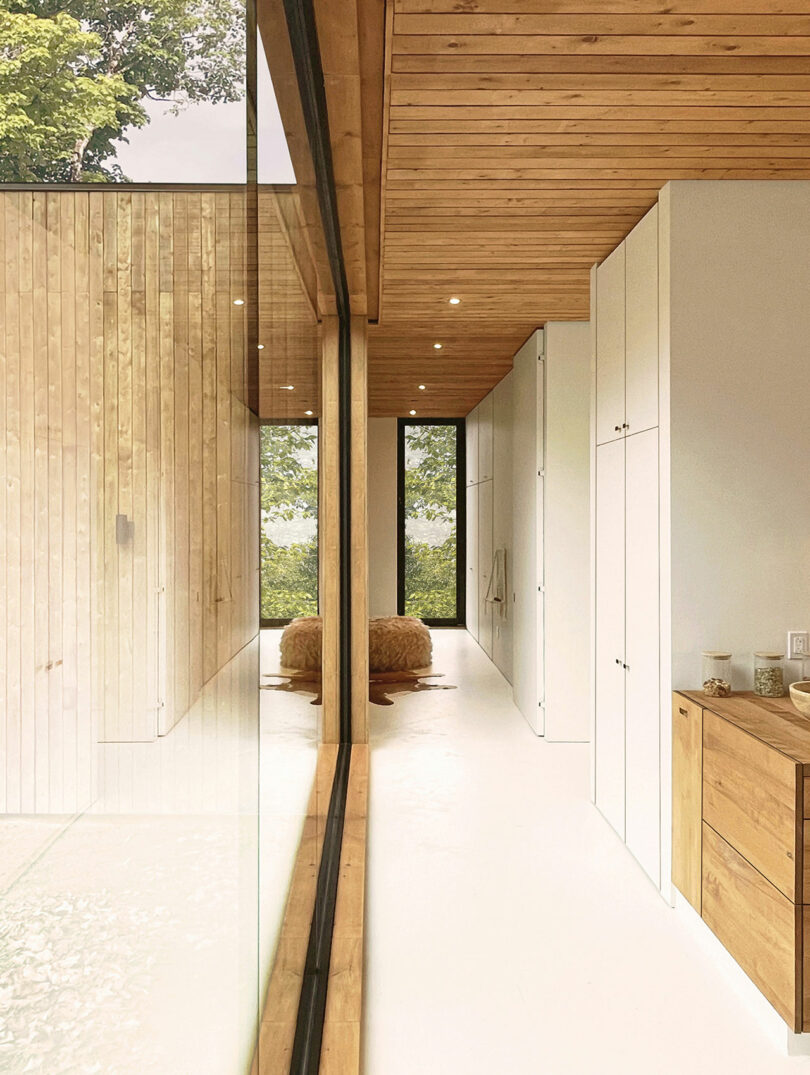 Modern interior hallway with white walls, wooden ceiling, built-in wardrobes and large floor-to-ceiling windows overlooking trees; There are two brown stools at the end of the hall.