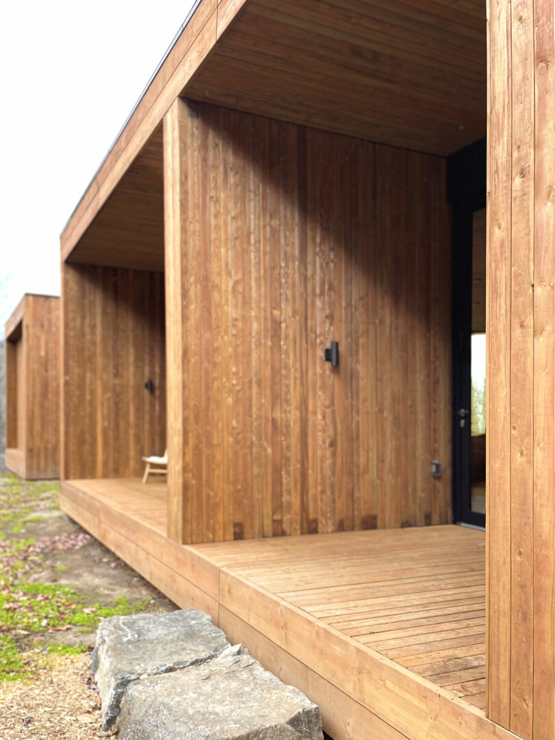 A modern building with vertical wooden cladding and a covered veranda with a single chair and large stones next to the wooden terrace.