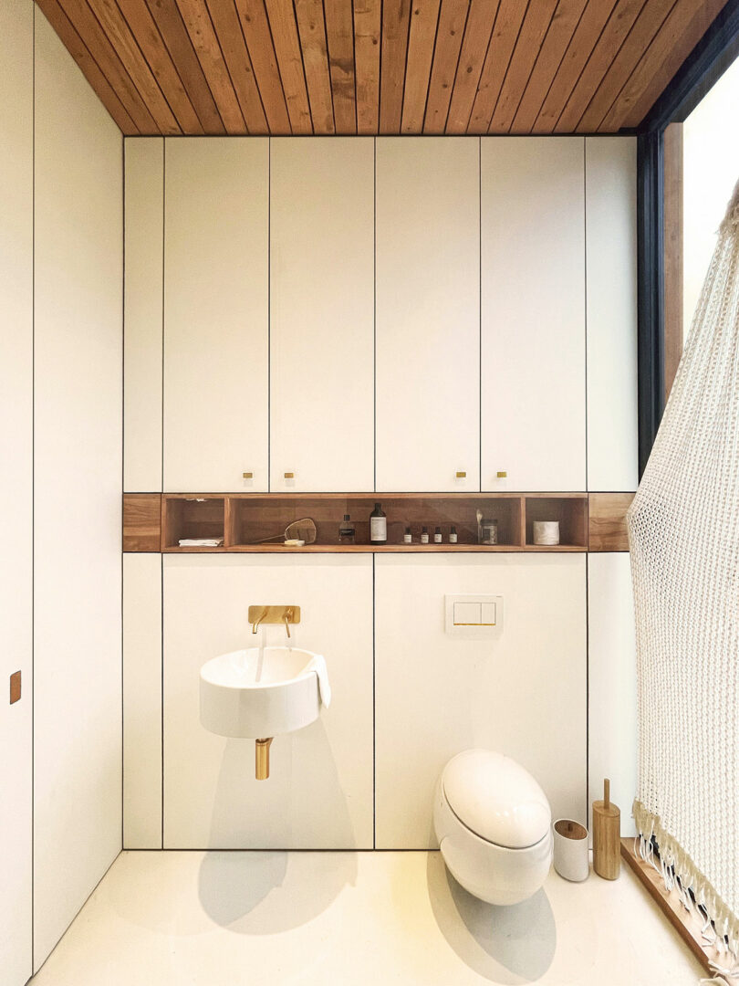 Modern, minimalist bathroom with white cabinets, a round wall-mounted sink, an oval toilet, wooden accents and a partially visible hanging chair on the right.