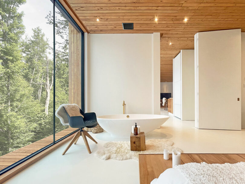 Modern bathroom with freestanding bathtub, wooden stool, black chair and floor-to-ceiling window with a view of the trees; minimalist decor with white walls and ceiling.