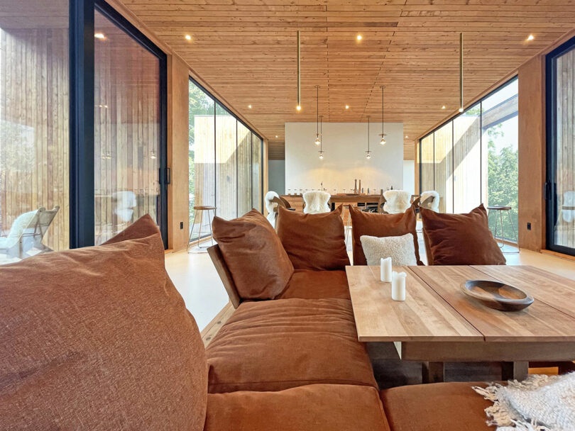 Modern living and dining area with large brown sofa set, wooden table and minimalist decor with floor-to-ceiling windows and wood-paneled ceilings and walls.