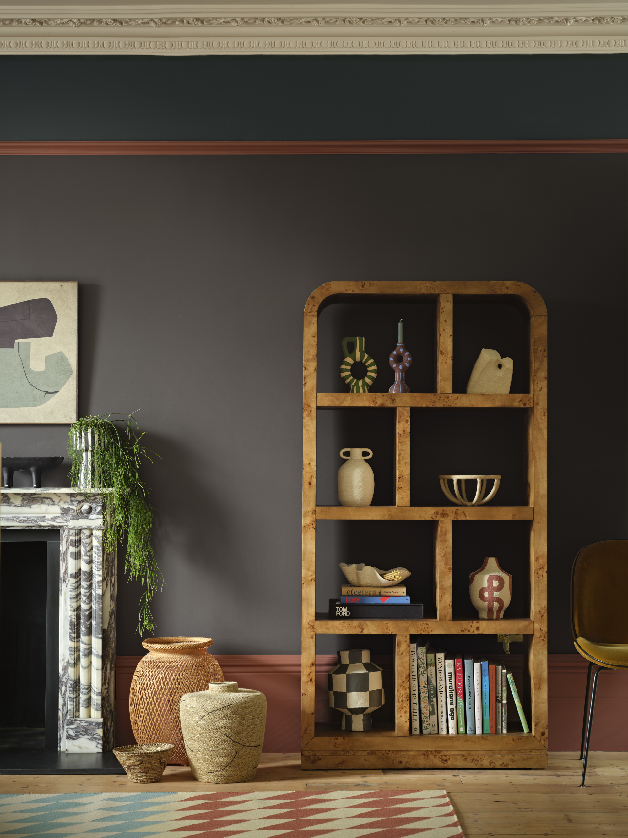 Image of a wall painted a dark purple-gray with coral trim and dark teal color blocking. There is a large burl wood shelf and a marble fireplace with flower pots in front of it.