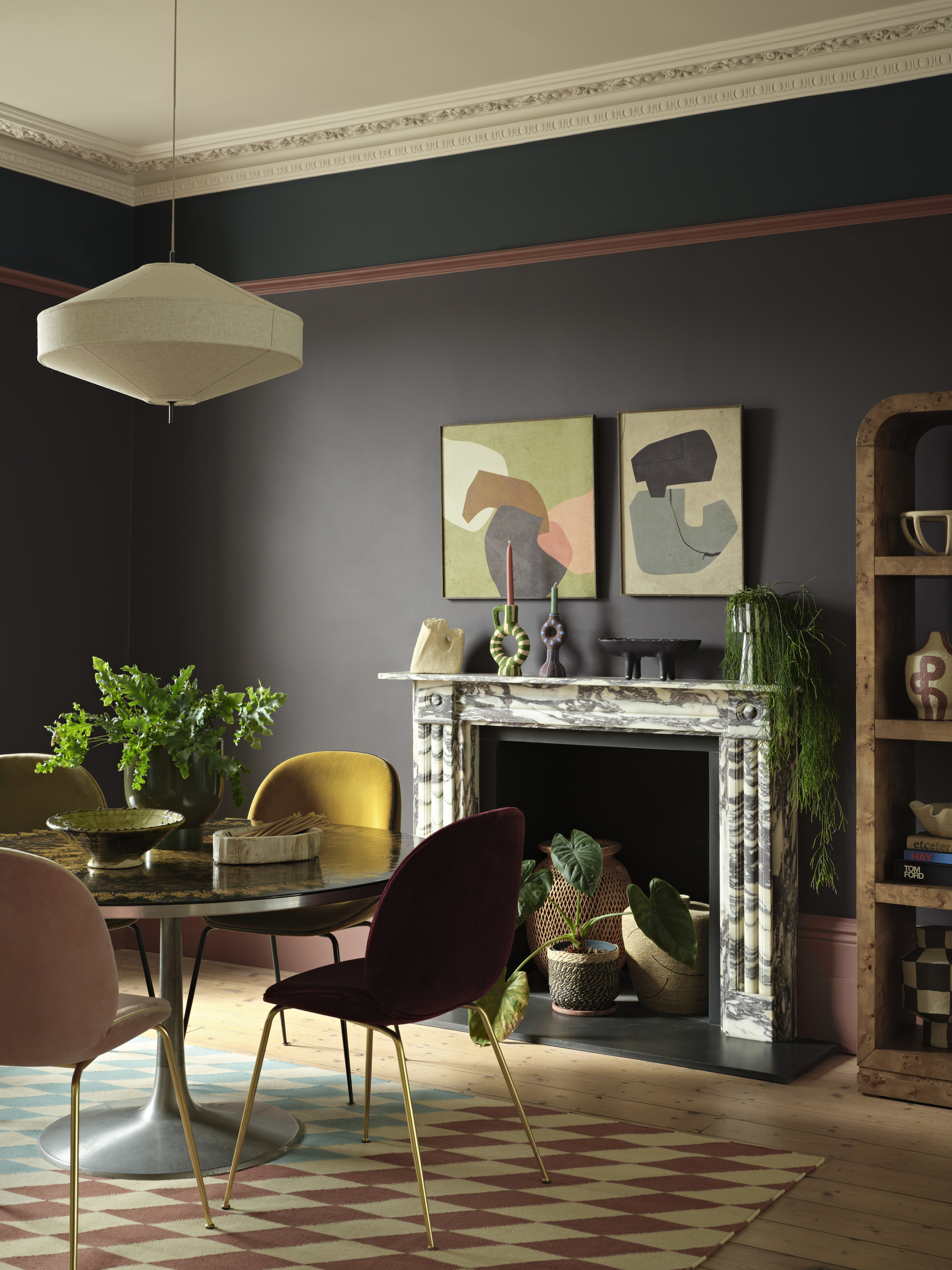 Image of a dining area in a room painted in a dark gray-purple shade with pink and teal trim. There is a marble fireplace with two paintings above it and a round dining table with jewel-toned seating.