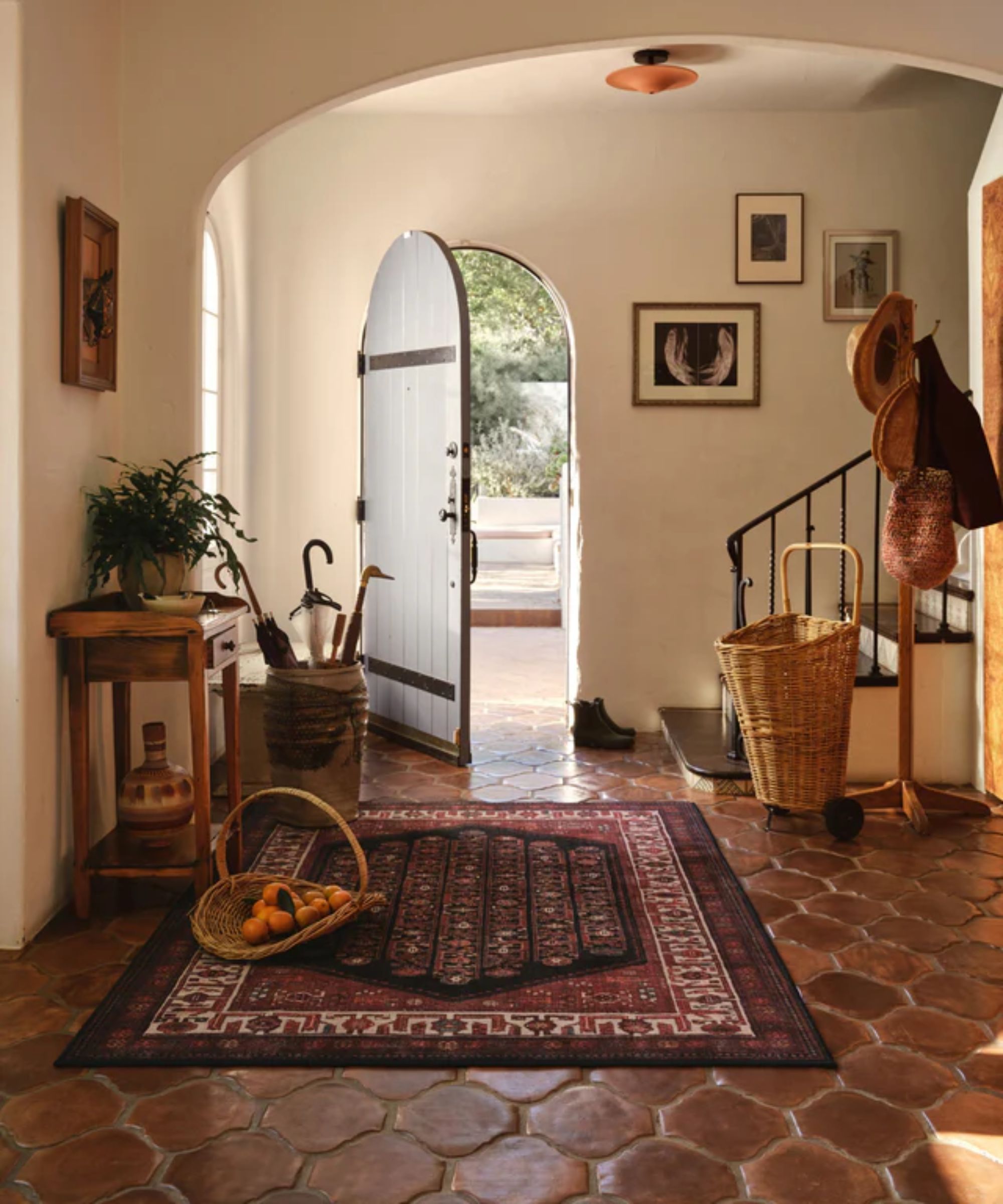Neutral entryway in a Spanish-style home with an arched front door and dark terracotta floors, styled with a large dark red rug, wooden table and vintage baskets
