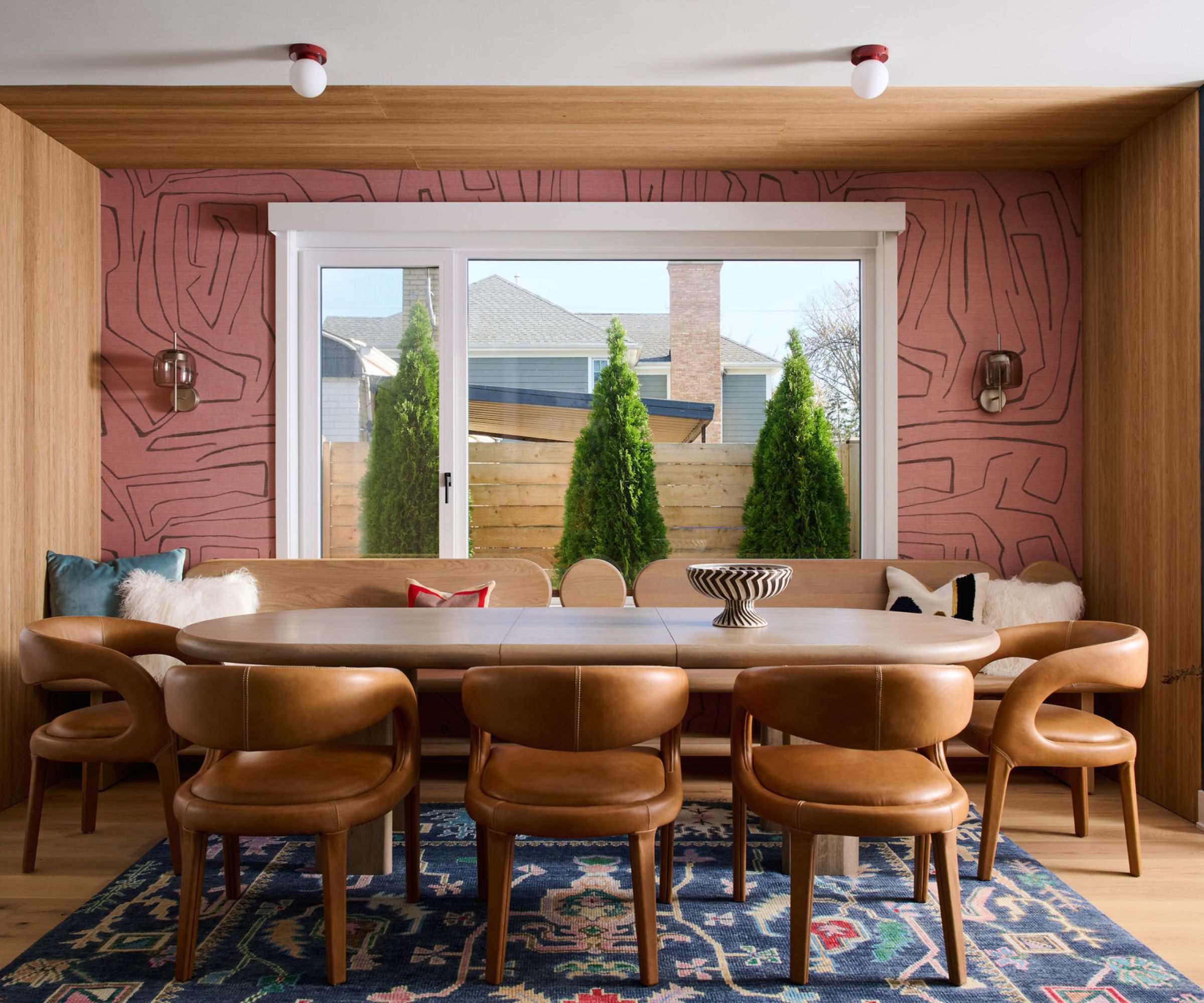 Dining room with pink abstract wallpaper, long curved wooden dining table, brown leather chairs, banquette and Oushak rug