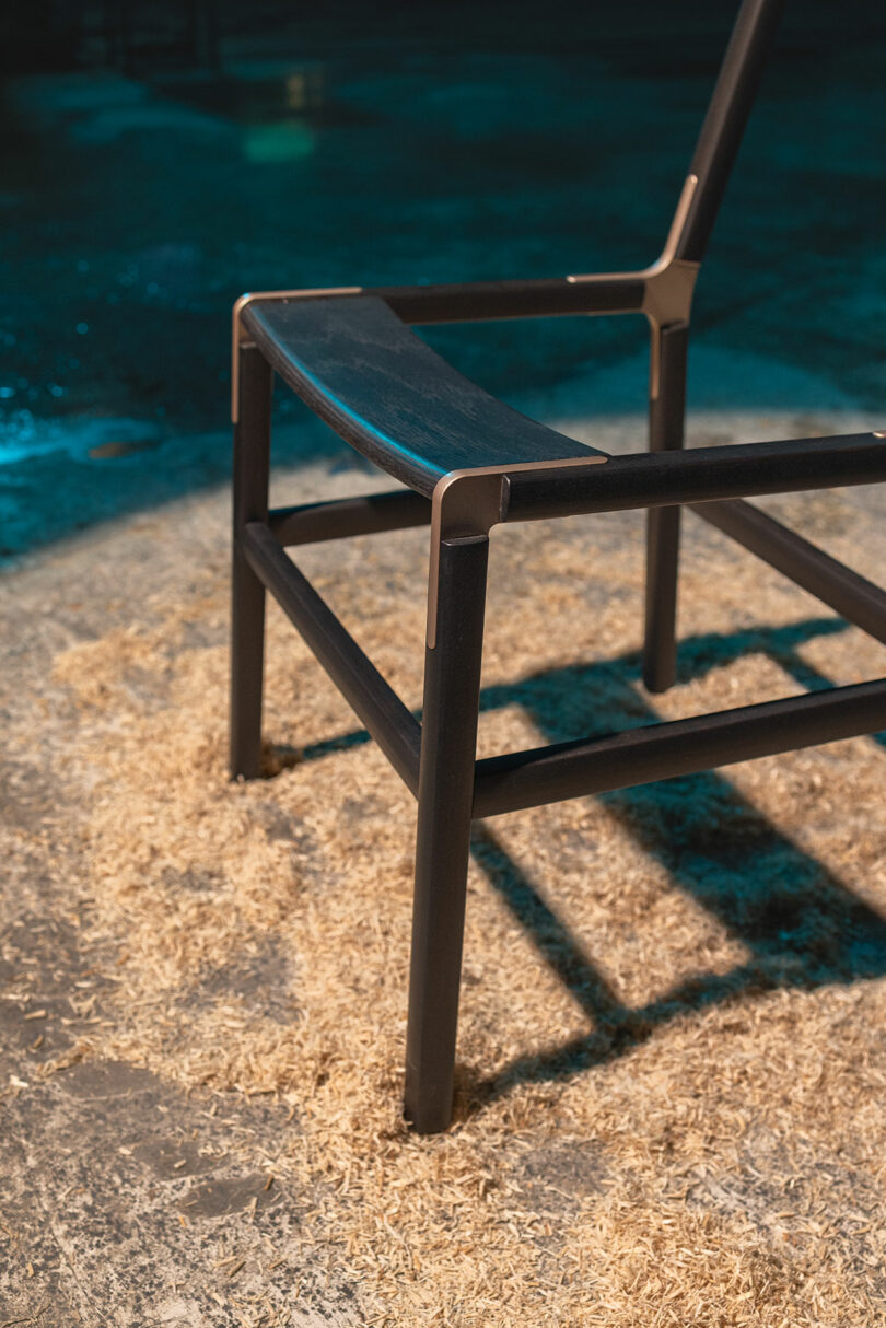 A modern black metal chair with a wooden seat stands on a surface covered in sawdust in atmospheric lighting.