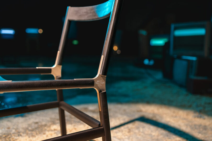 Close-up of a modern black chair with metal corner joints against a blurred background with blue and teal lighting.