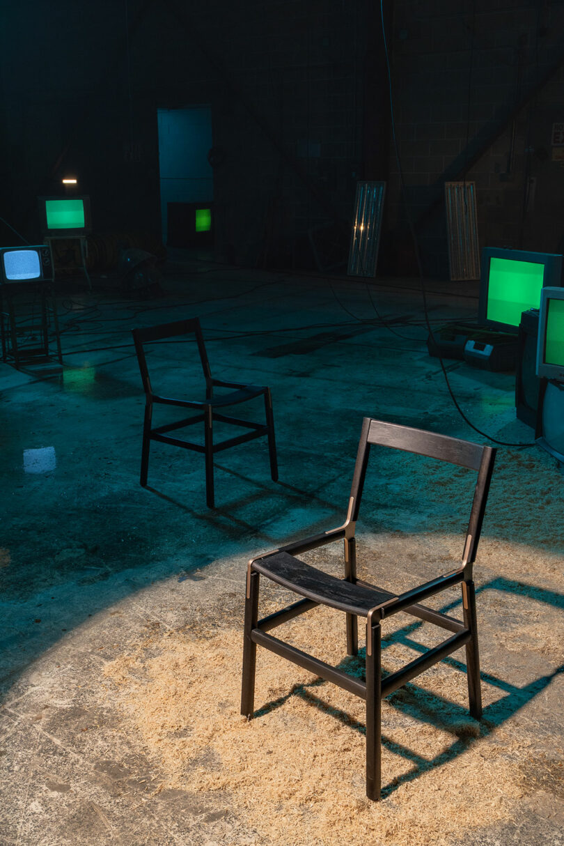 A black wooden chair stands in the spotlight on a rough concrete floor, in the background two other chairs, green-lit television screens and scattered sawdust.