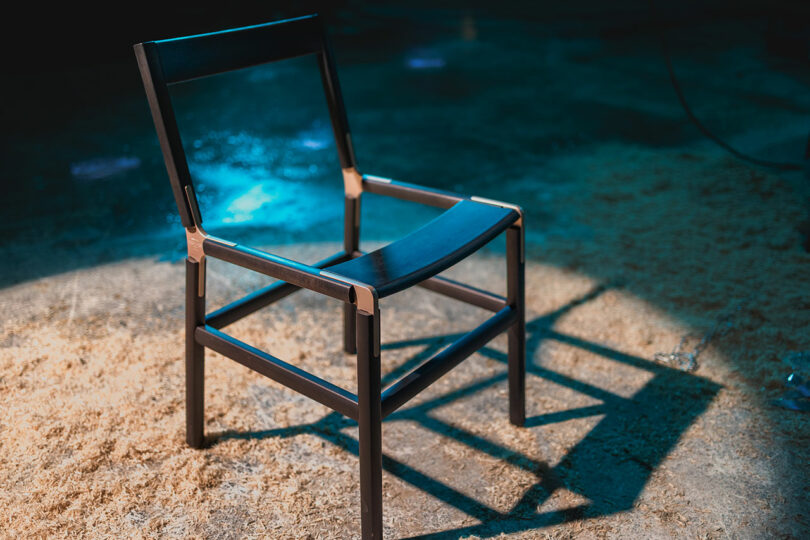 A modern black wooden chair with a minimalist design stands on a textured floor covered with sawdust under blue lighting.