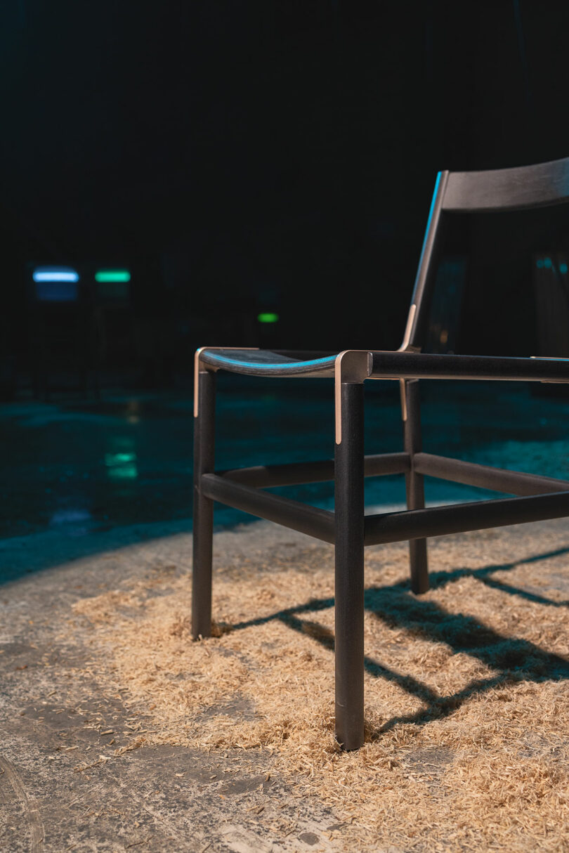 A black wooden chair stands on a concrete floor covered in sawdust, in low lighting with a dark blurry background.