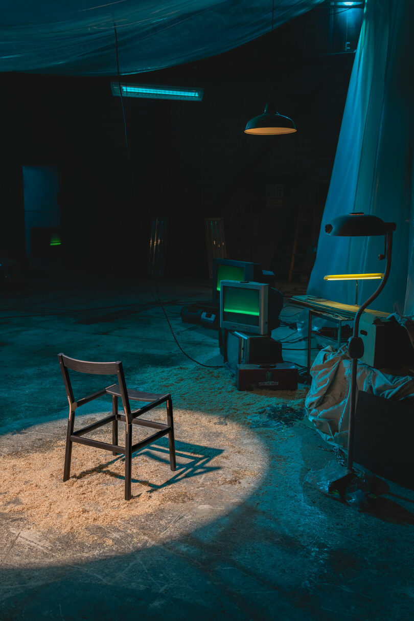 A single wooden chair is illuminated by a spotlight in a dimly lit industrial room with scattered old electronics and plastic sheeting.