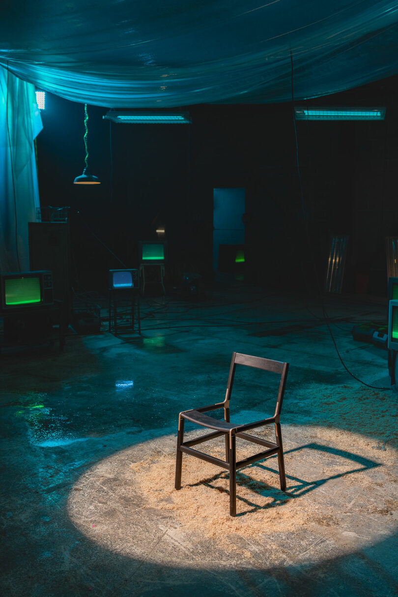 A single wooden chair sits under a spotlight in a dark industrial room with scattered sawdust and several old televisions emitting green light.