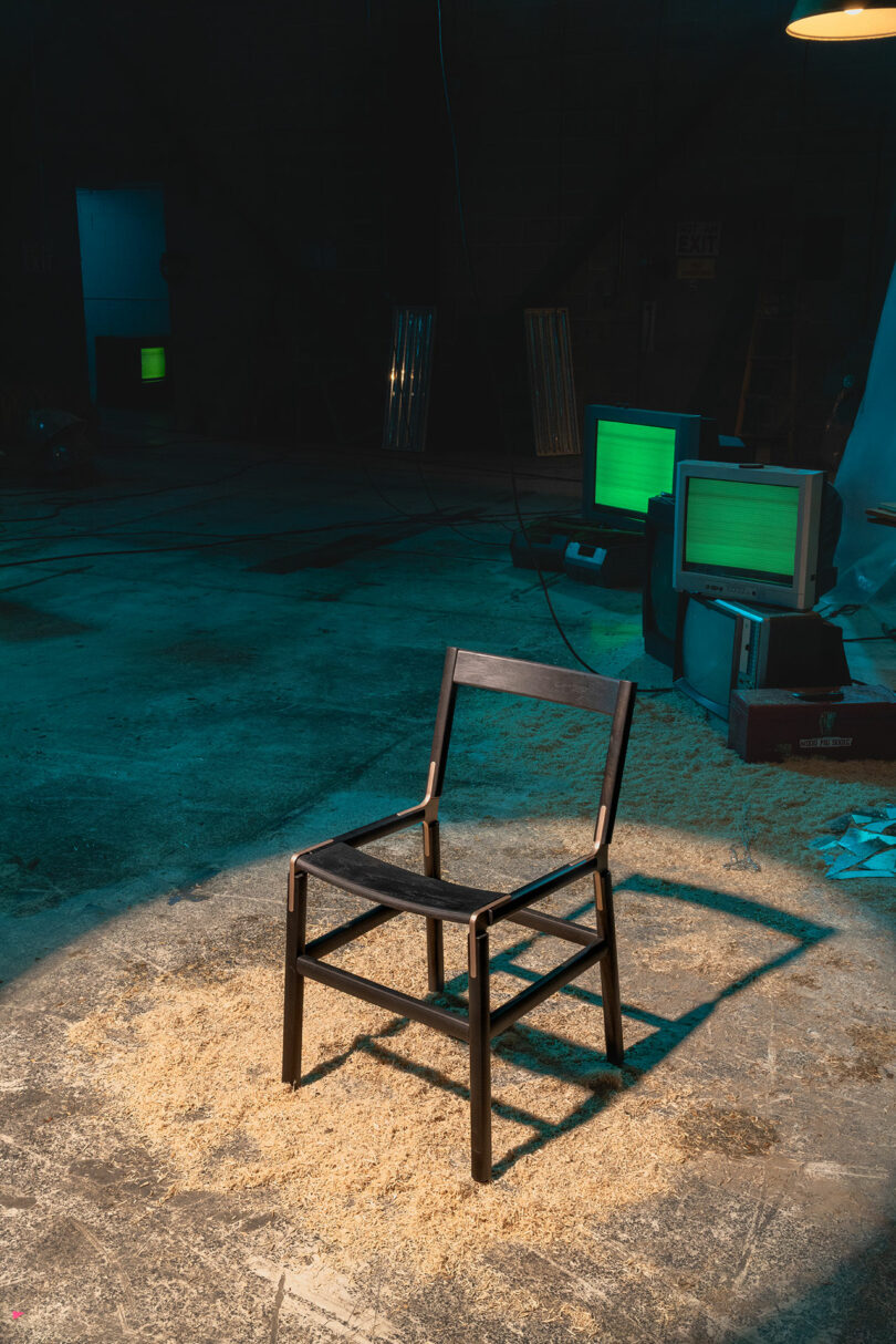 A wooden chair sits on a sawdust-covered floor in a dimly lit room with old computer monitors in the background.