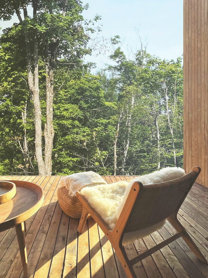 A wooden deck with lounge chair, sheepskin throw, round ottoman and side table overlooks a lush, green forest.