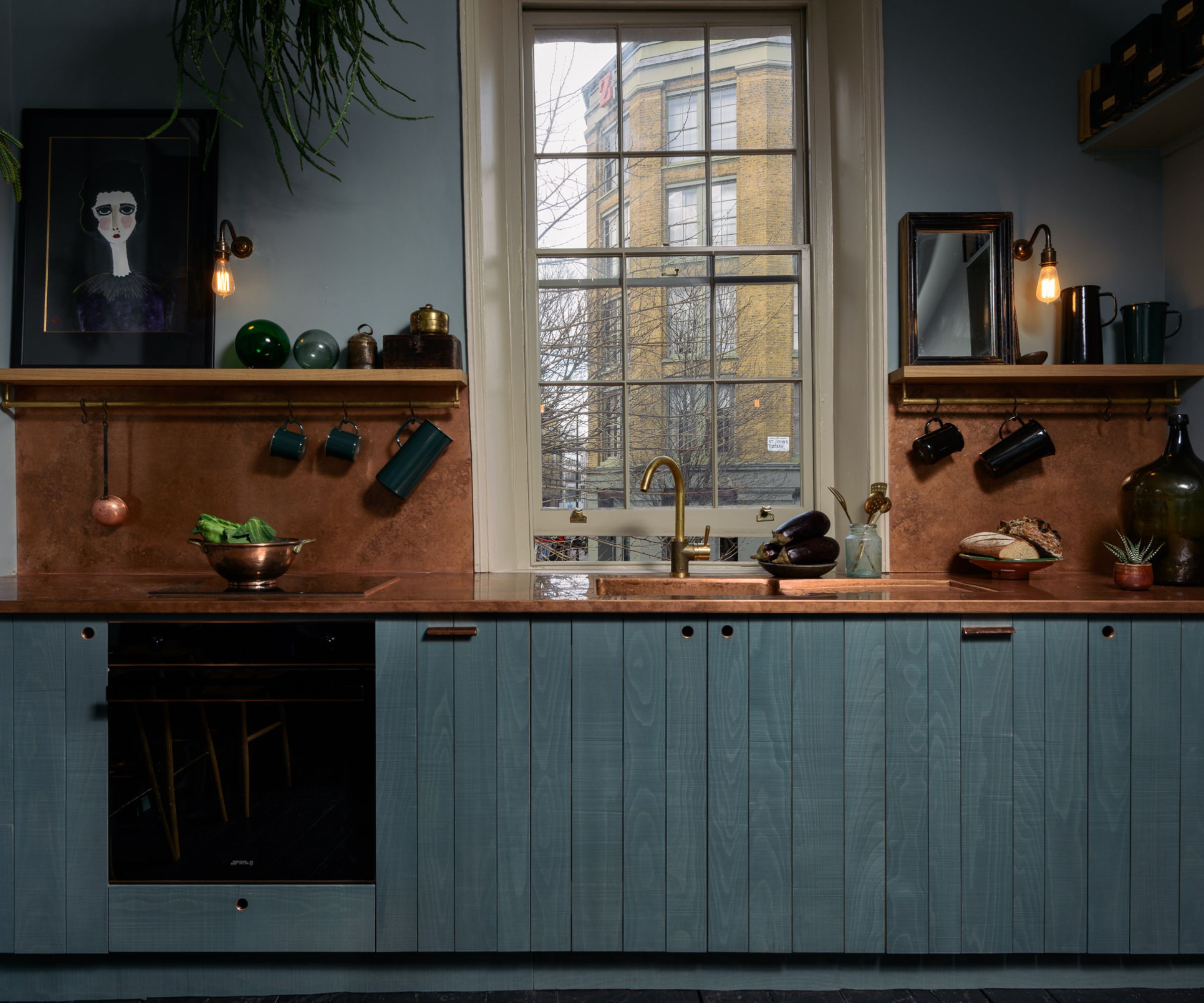 A single-wall kitchen with blue cabinets, copper countertops, and open shelves filled with art and kitchenware