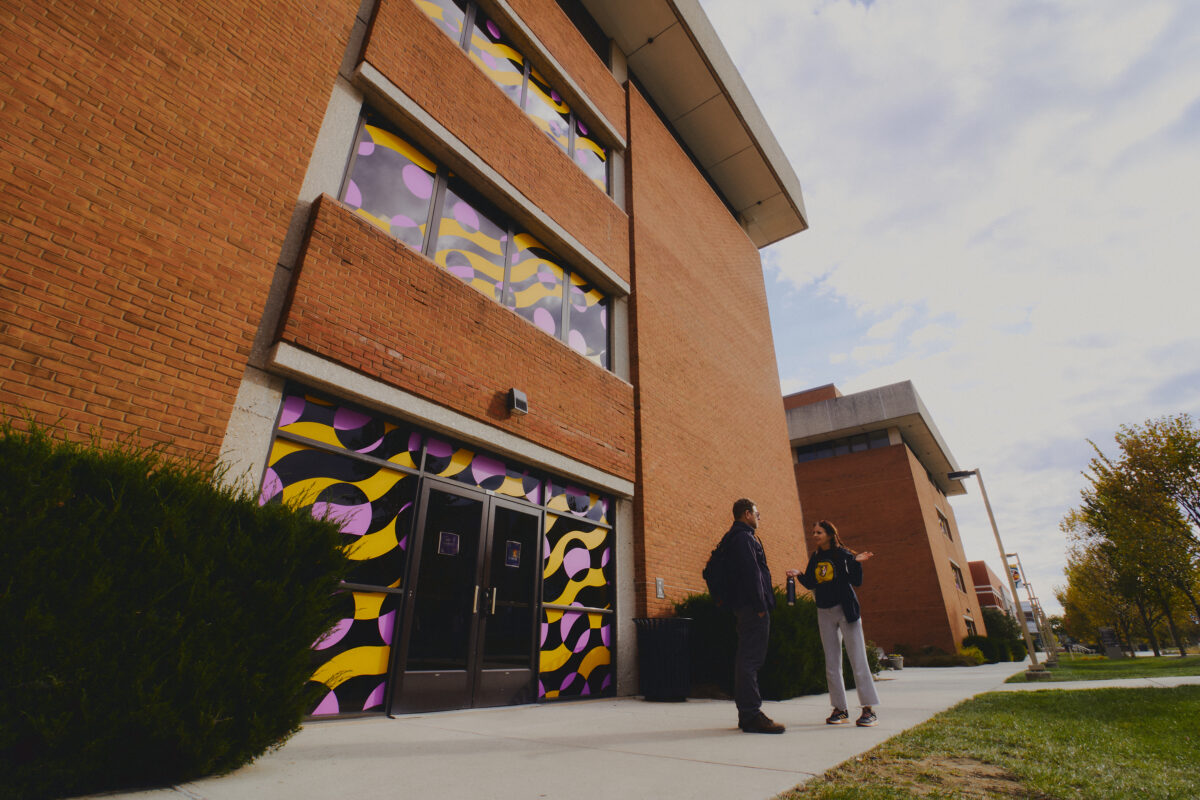 Two people talk in front of a brick building with colorful abstract window designs in yellow, pink and black.