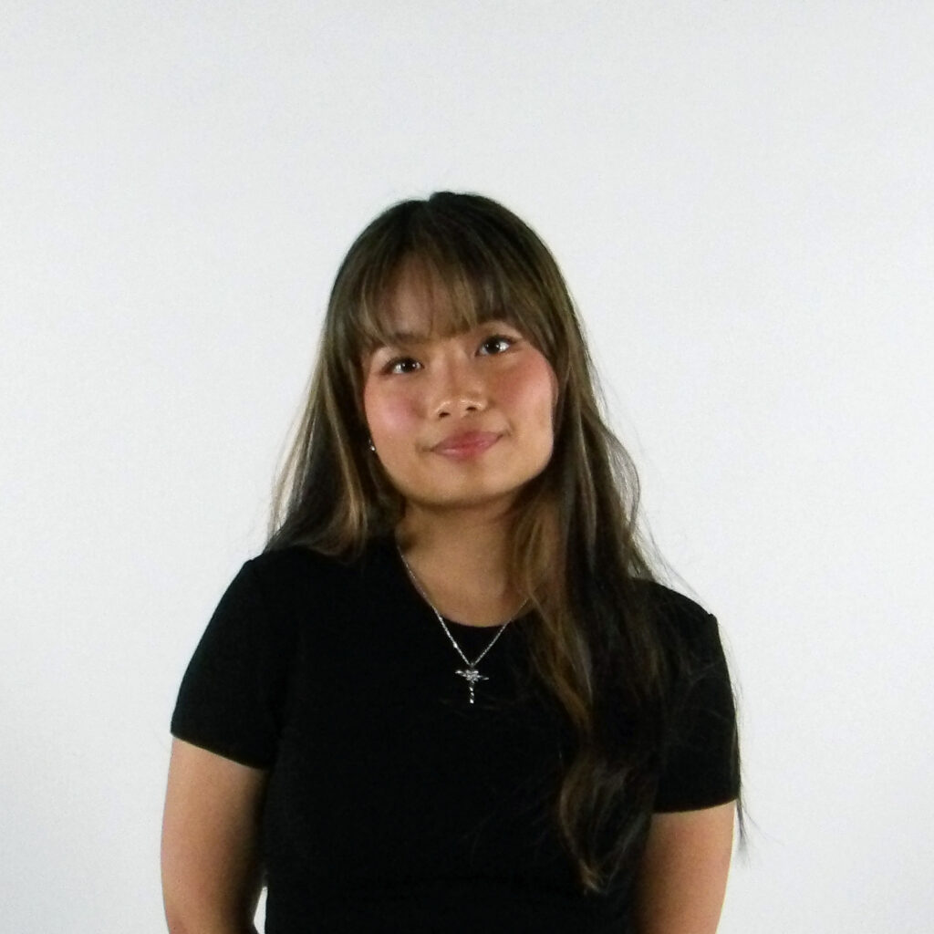 A person with long dark hair, a black top and a necklace stands in front of a white background.