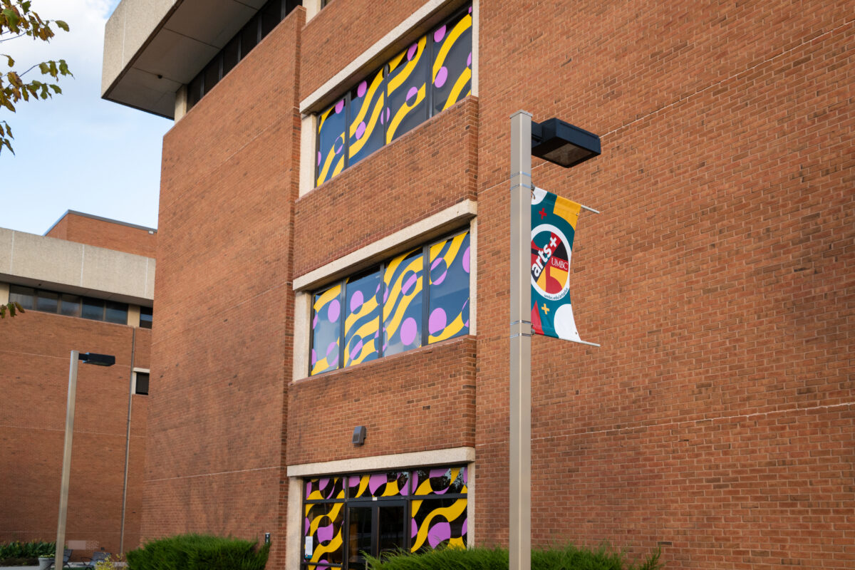 A brick building with windows decorated in a yellow and purple abstract pattern and a nearby pole with a colorful