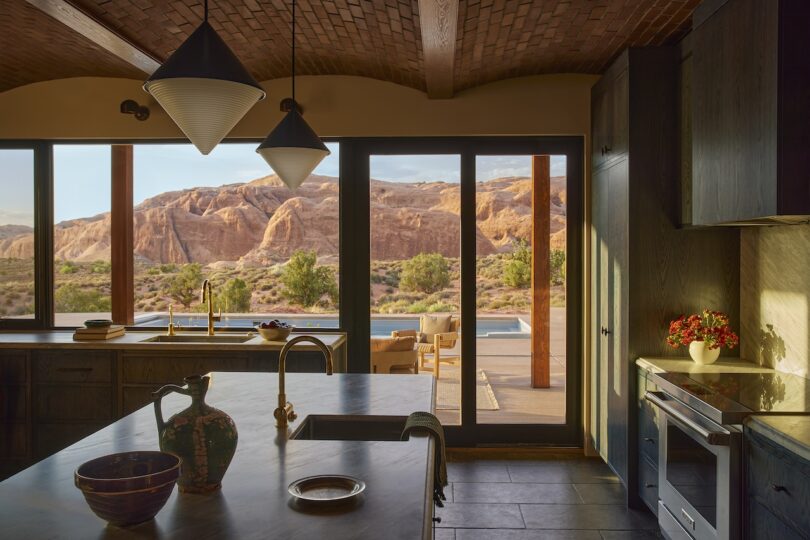 Modern kitchen with dark cabinets and pendant lights, large windows and doors leading to a patio with a pool and scenic views of the red rock desert cliffs.