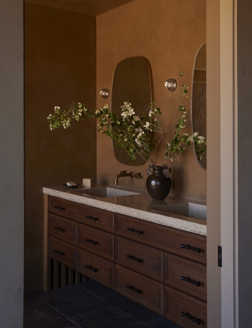 A bathroom vanity with a marble countertop, two sinks, wooden drawers, two mirrors, a dark vase with branches and warm brown walls.