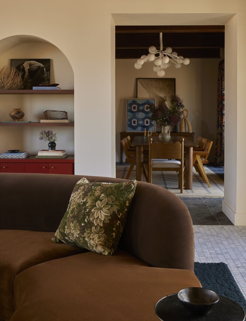 A living room with a brown curved sofa, a floral pillow, built-in shelves and a view into a dining area with a wooden table, chairs, a modern chandelier and framed artwork.