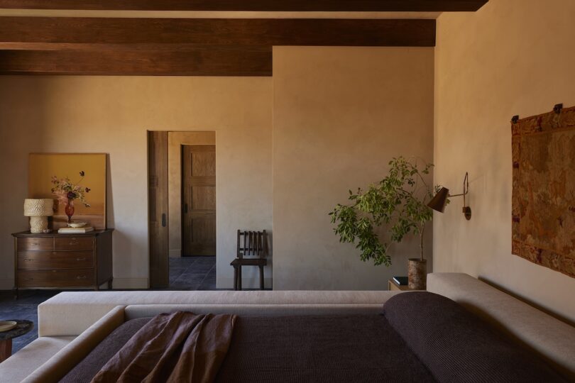 A bedroom with beige walls, beamed ceilings, a bed with dark linens, a dresser, a chair, a potted plant and wall art. Warm natural light penetrates the room.