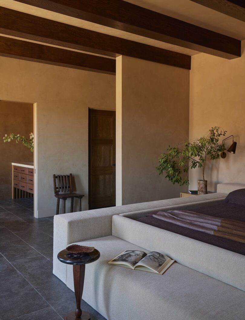 Minimalist bedroom with exposed beam ceilings, neutral-toned walls, low bed, potted plants, open book on a side table and a view into an adjacent room with a wooden bench.
