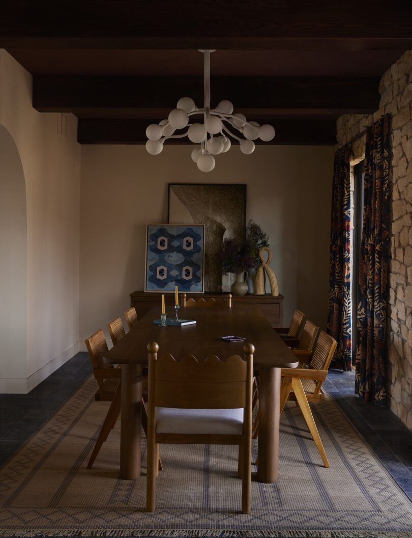 A dining room with a wooden table and six chairs, a modern chandelier, a sideboard with decor, framed art and patterned curtains in neutral and earthy tones.