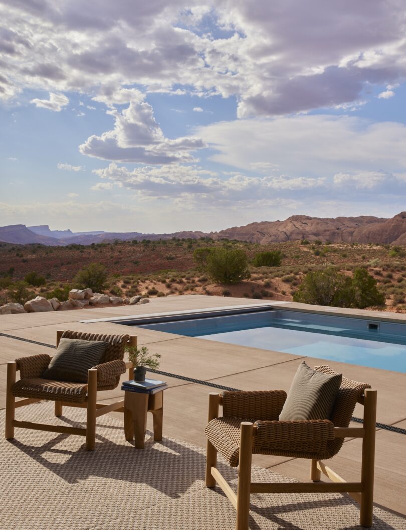 Two wicker chairs with cushions and a small table stand by a swimming pool and overlook a desert landscape with hills and a cloudy sky in the background.
